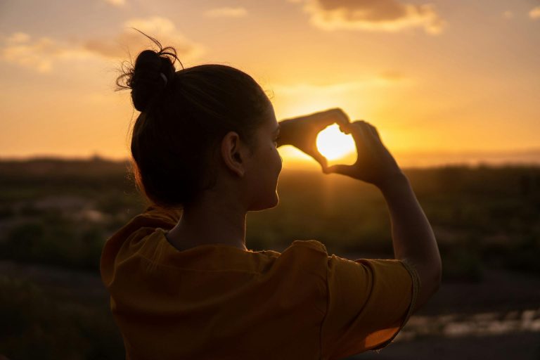 Amor propio como base de la felicidad: mujer formando un corazón al atardecer.