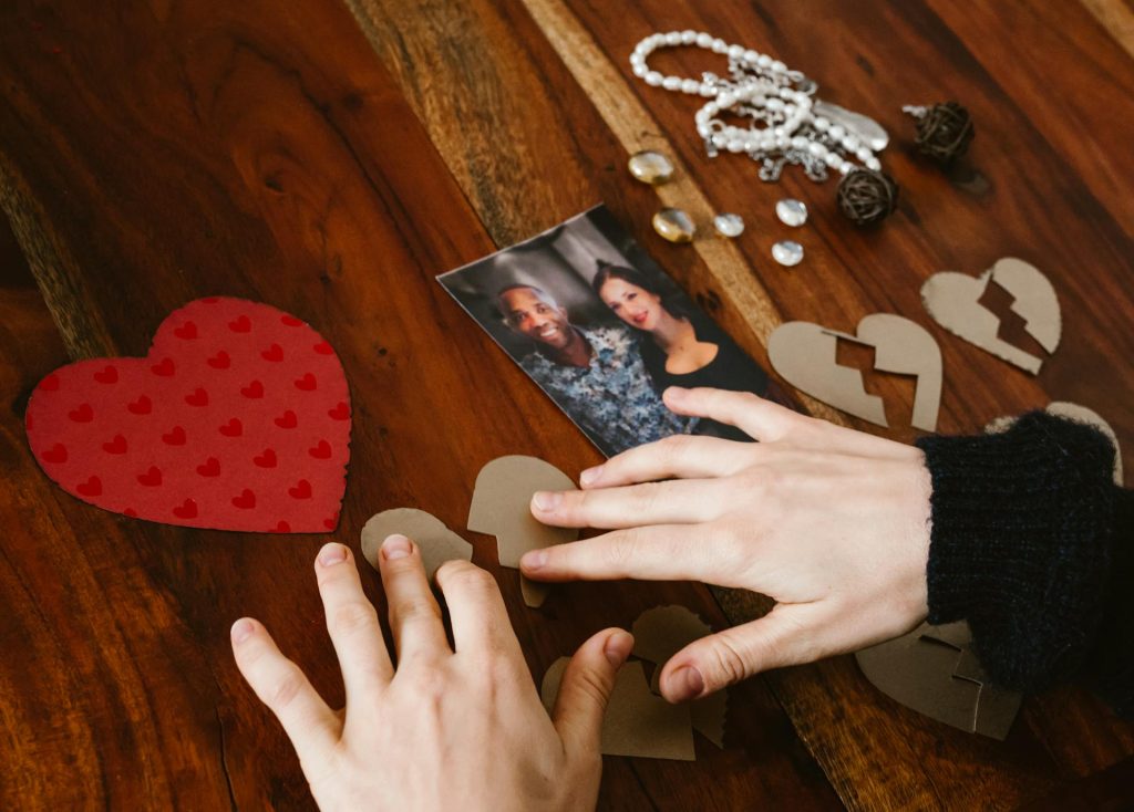 Persona uniendo piezas de un corazón roto de cartón sobre una mesa de madera junto a una fotografía de pareja, simbolizando la ruptura por repetición de patrones en las relaciones de pareja.