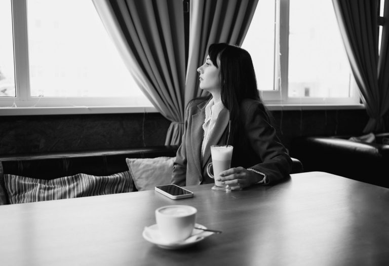 Mujer sentada sola en una cafetería mirando por la ventana mientras sostiene una bebida de café, representando la hiperindependencia, la dificultad para conectar emocionalmente y los desafíos de las mujeres exitosas en sus relaciones de pareja.
